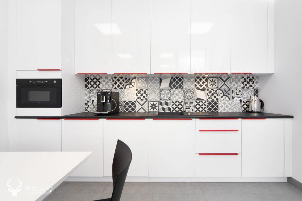 Biała, nowoczesna kuchnia z czarnym blatem i czerwonymi akcentami, mozaika z wzorzystych płytek nad blatem, ekspres do kawy i czajnik.