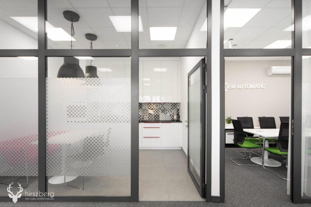 Przeszklona ścianka działowa w biurze, widok na salę konferencyjną z zielonymi krzesłami i aneks kuchenny z mozaiką nad blatem.