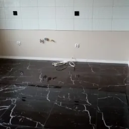 Ciemna, błyszcząca podłoga z płytek imitujących marmur, świeżo położona, z widocznymi przewodami elektrycznymi i przygotowanym miejscem na armaturę, białe płytki ścienne na górze, częściowo ułożone...