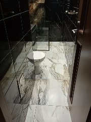 Wnętrze łazienki z toaletą, wykończone czarnymi płytkami na ścianach i marmurowymi płytkami na podłodze. Widoczny fragment kabiny prysznicowej i drzwi. Oświetlenie górne tworzy refleksy...