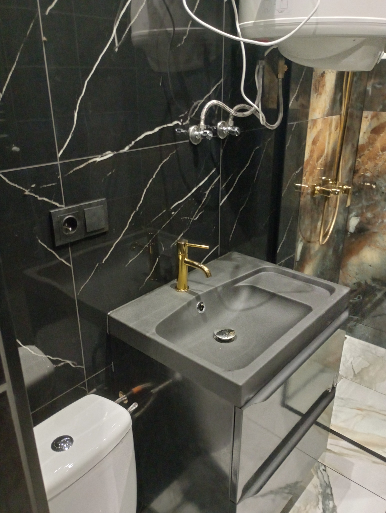 Szara umywalka z szafką i złotą baterią na tle czarnych płytek imitujących marmur, obok toaleta i złota deszczownica.