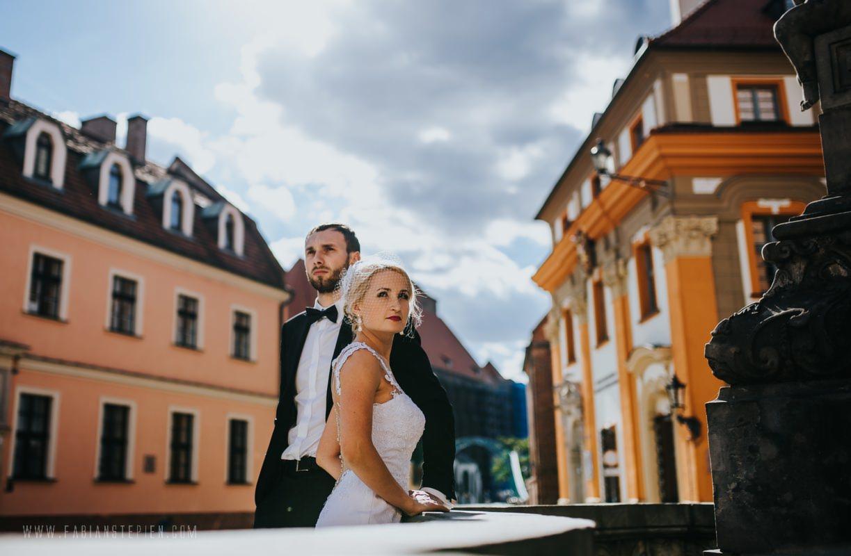 Para młoda w eleganckich strojach pozuje na tle zabytkowych kamienic w słoneczny dzień, panna młoda z czerwoną szminką patrzy w obiektyw.