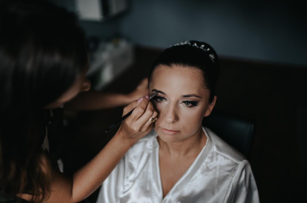 Kobieta w satynowym szlafroku, z tiarą we włosach, podczas wykonywania makijażu brwi przez wizażystkę z pierścionkiem na palcu.