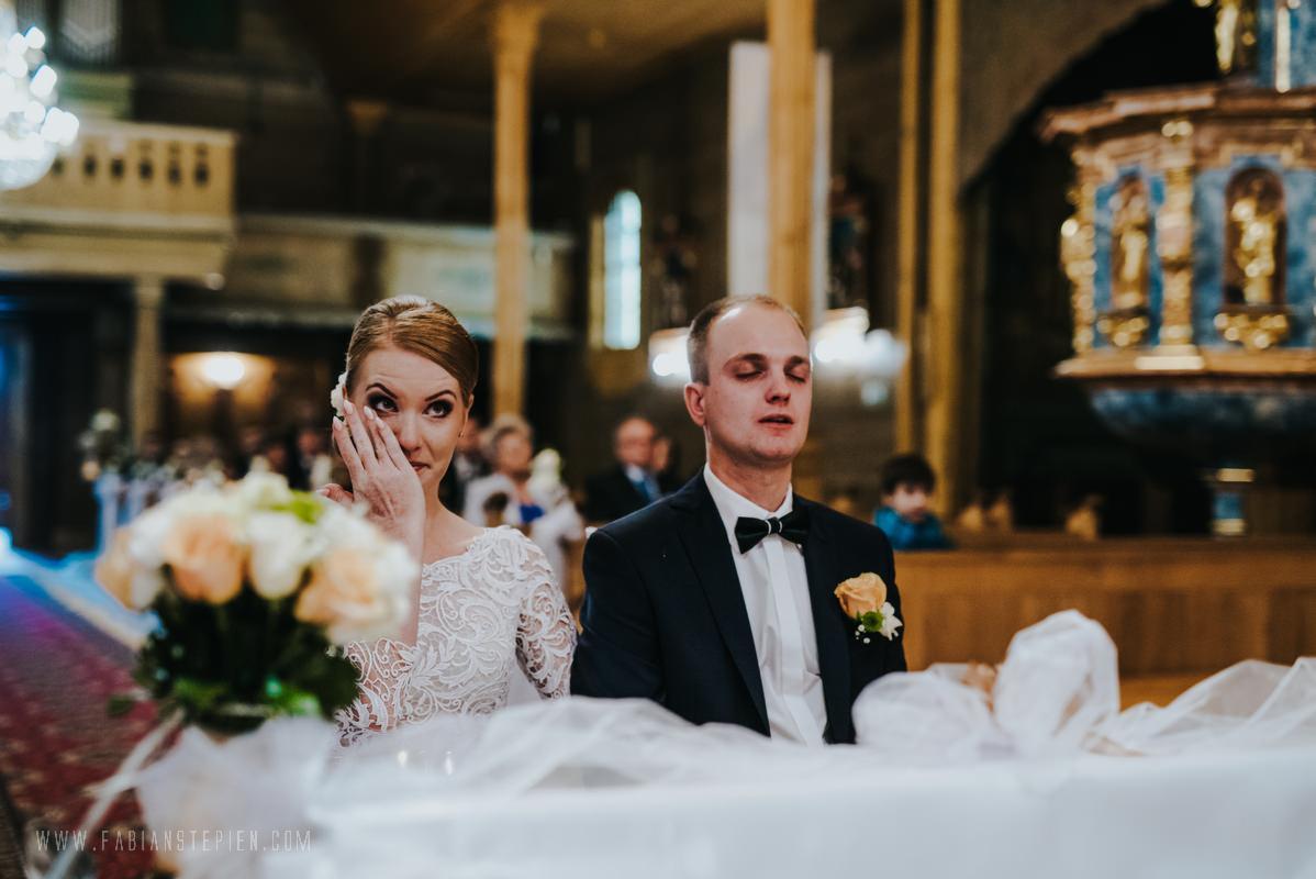 Panna młoda ociera łzę wzruszenia podczas ceremonii ślubnej w kościele, obok stoi zamyślony pan młody w ciemnym garniturze z muszką i butonierką z jasną różą, w tle widać ołtarz i gości.