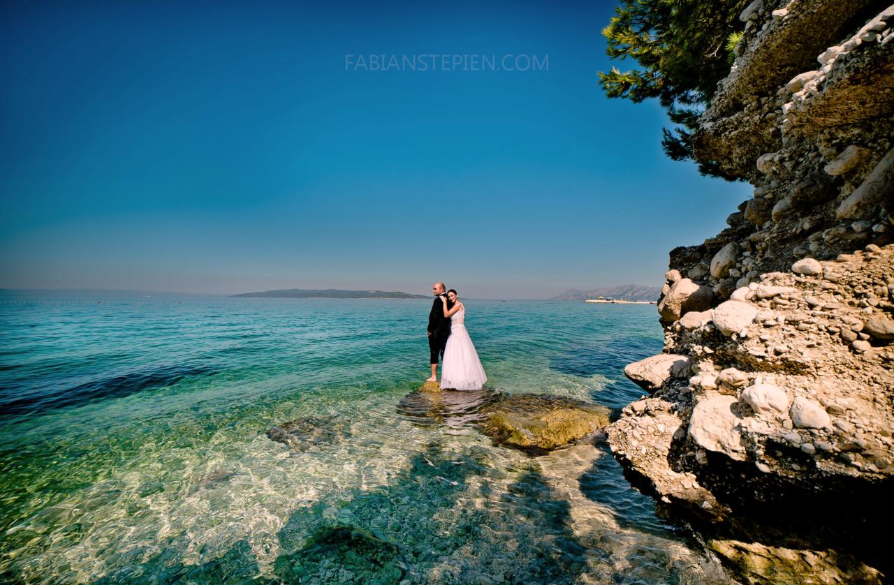 Para młoda w Panki stoi na skałach w krystalicznie czystej wodzie morza, z skalistym brzegiem po prawej stronie i błękitnym niebem nad nimi.