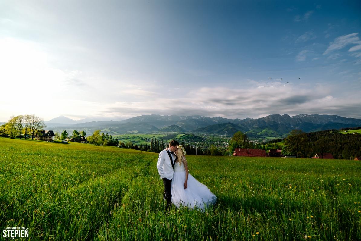 Para młoda w objęciach na rozległej łące z widokiem na góry w tle, ona w białej sukni z wiankiem, on w białym smokingu, ujęcie z dołu z szerokim kadrem.