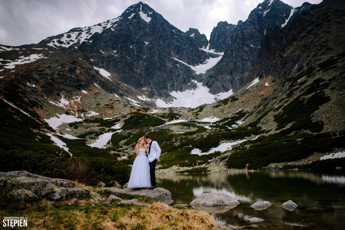 Para młoda w górach nad jeziorem, panna młoda w białej sukni i wianku, pan młody w białej marynarce, w tle skaliste szczyty z resztkami śniegu.