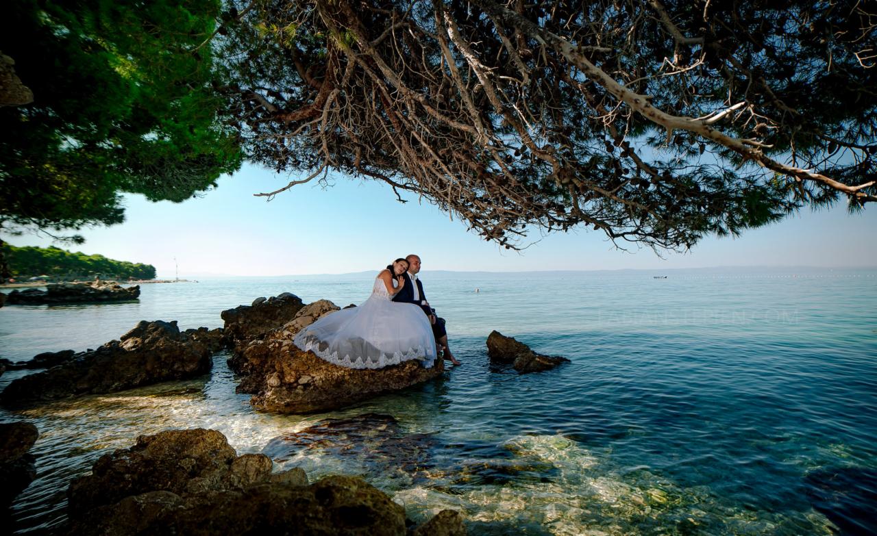 Para młoda siedzi na skałach nad brzegiem morza, z widokiem na horyzont i gałęzie drzew dające cień.