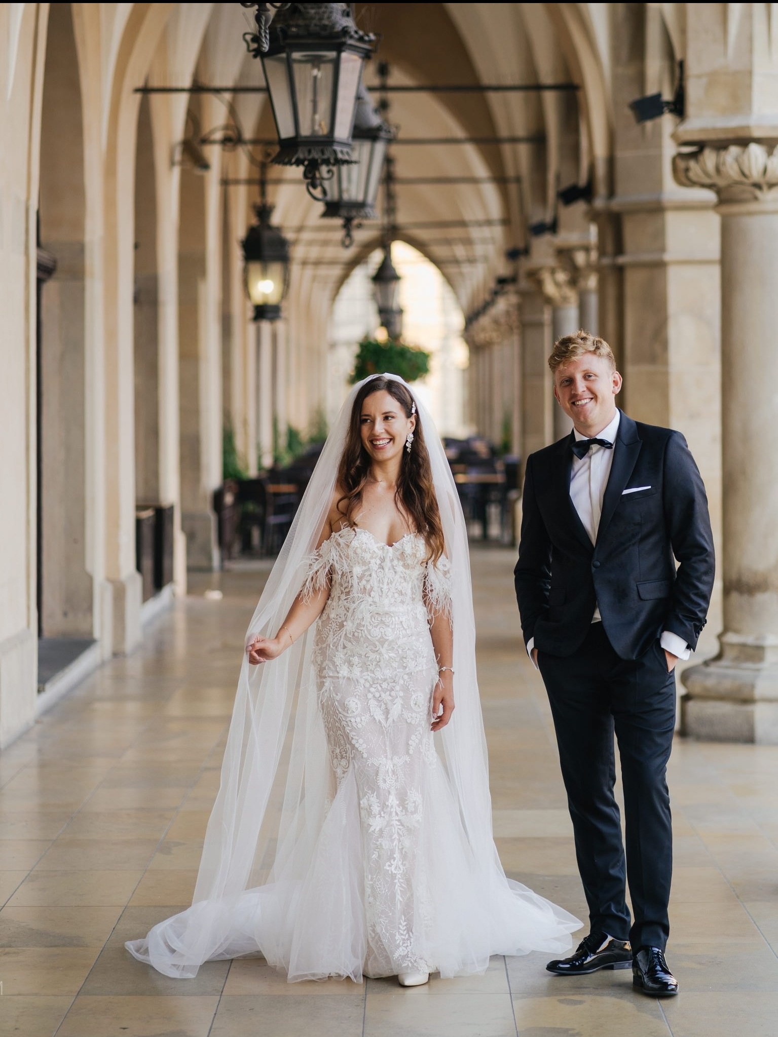 Uśmiechnięta para młoda w eleganckich strojach pozuje na tle kolumnady z latarniami. Panna młoda w koronkowej sukni z welonem, pan młody w czarnym smokingu.