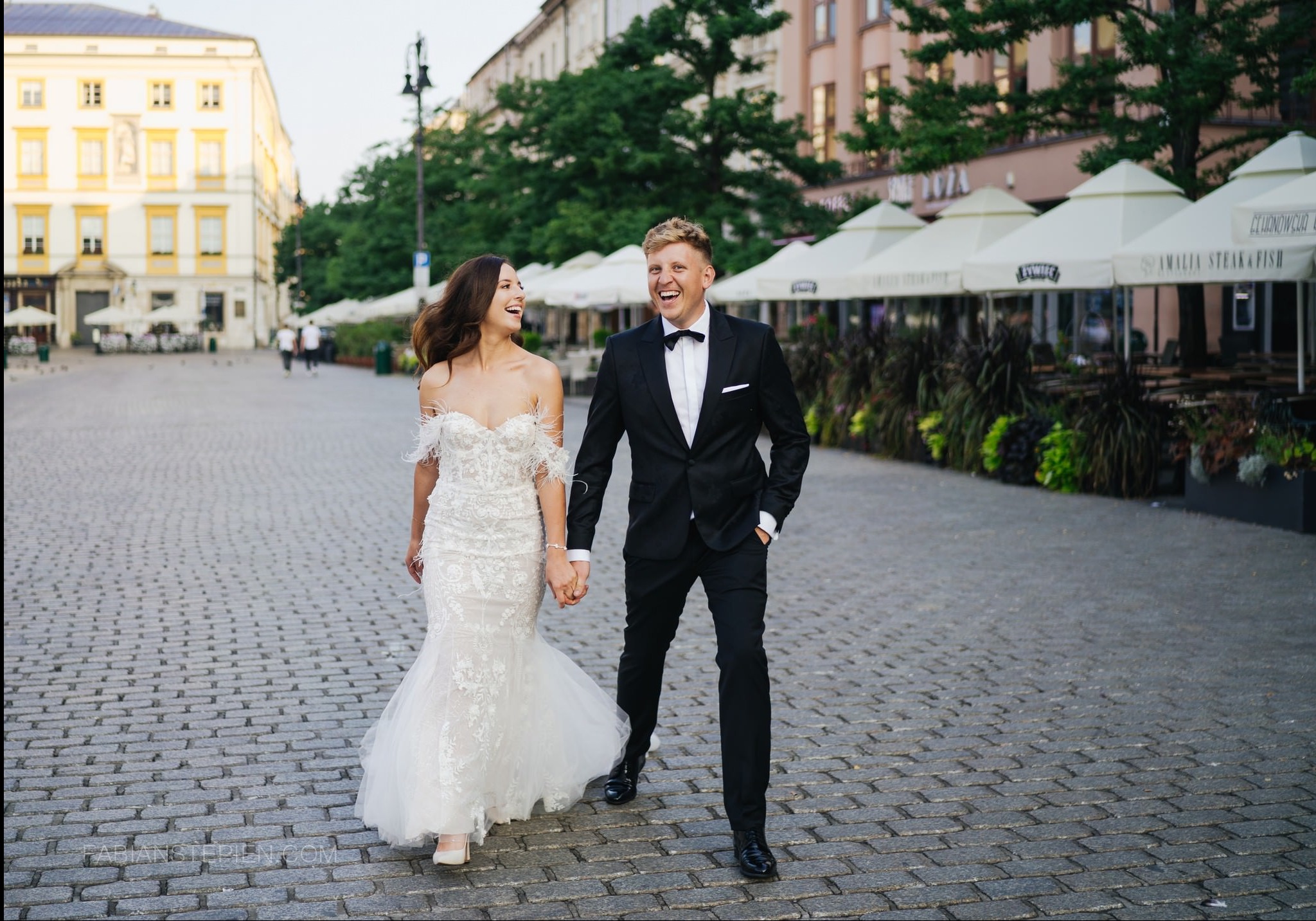Uśmiechnięta para młoda w eleganckich strojach spaceruje po brukowanej ulicy, w tle widoczne budynki i ogródki restauracyjne z parasolami.