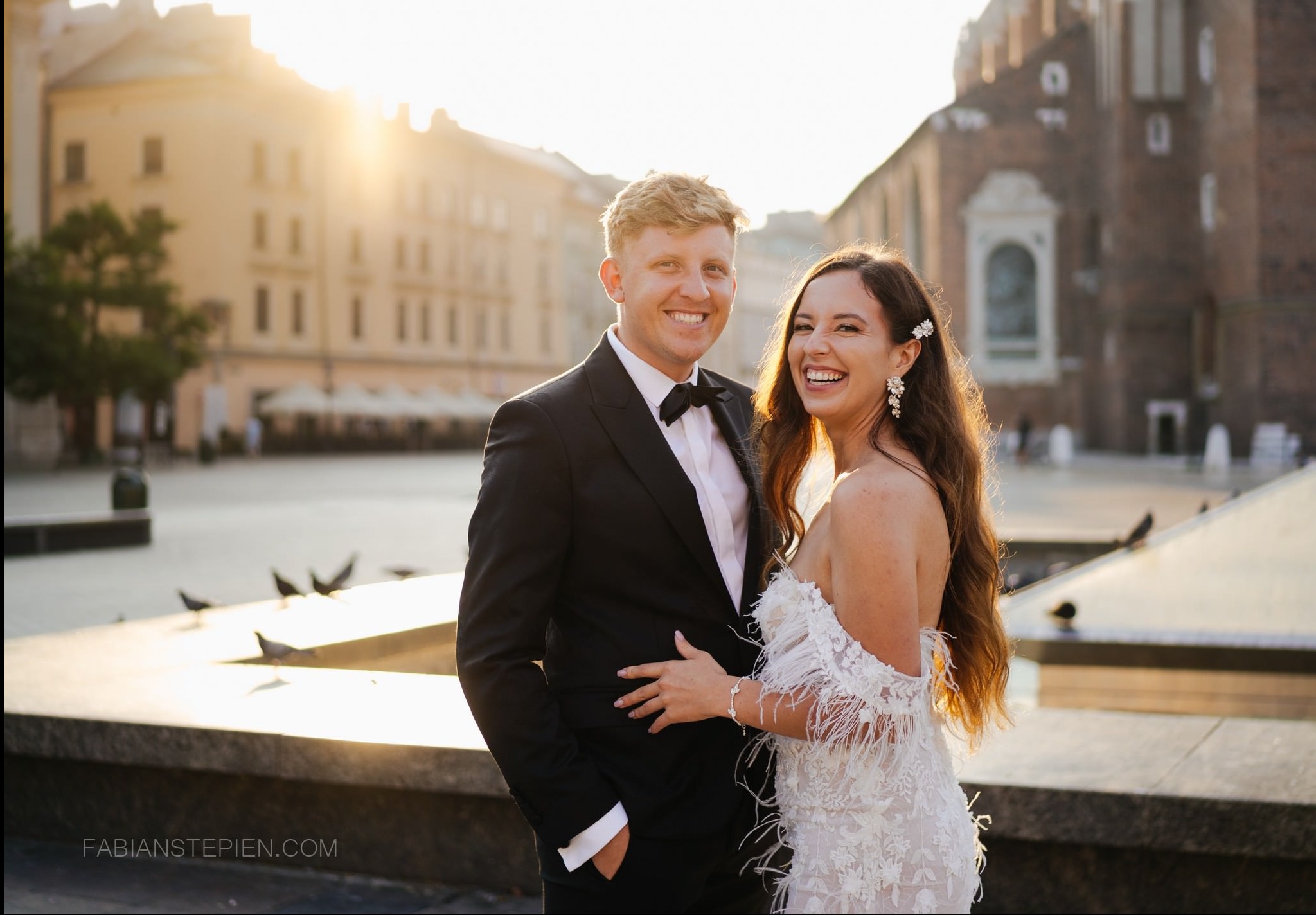 Uśmiechnięta para młoda w eleganckich strojach na tle Rynku Głównego w Krakowie, w promieniach zachodzącego słońca. Gołębie na pierwszym planie.