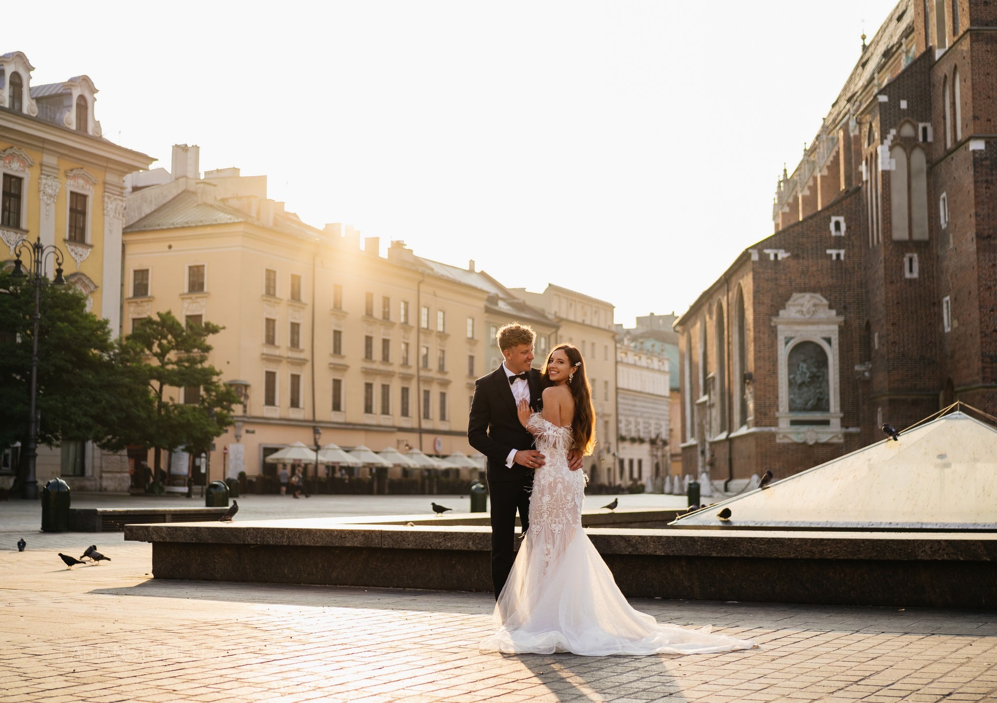 Para młoda w objęciach na tle Rynku Głównego w Krakowie, w promieniach zachodzącego słońca. Panna młoda w koronkowej sukni, pan młody w smokingu. Gołębie na bruku.