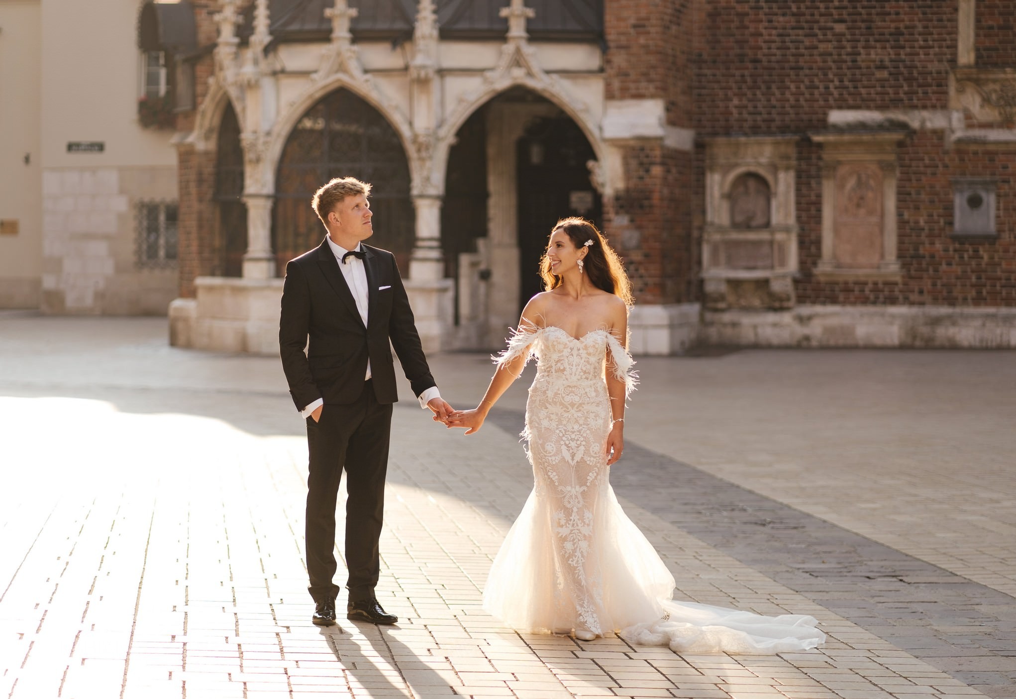 Para młoda trzymająca się za ręce na tle ceglanej architektury Krakowa, w promieniach zachodzącego słońca, ona w sukni z piórami, on w klasycznym smokingu.