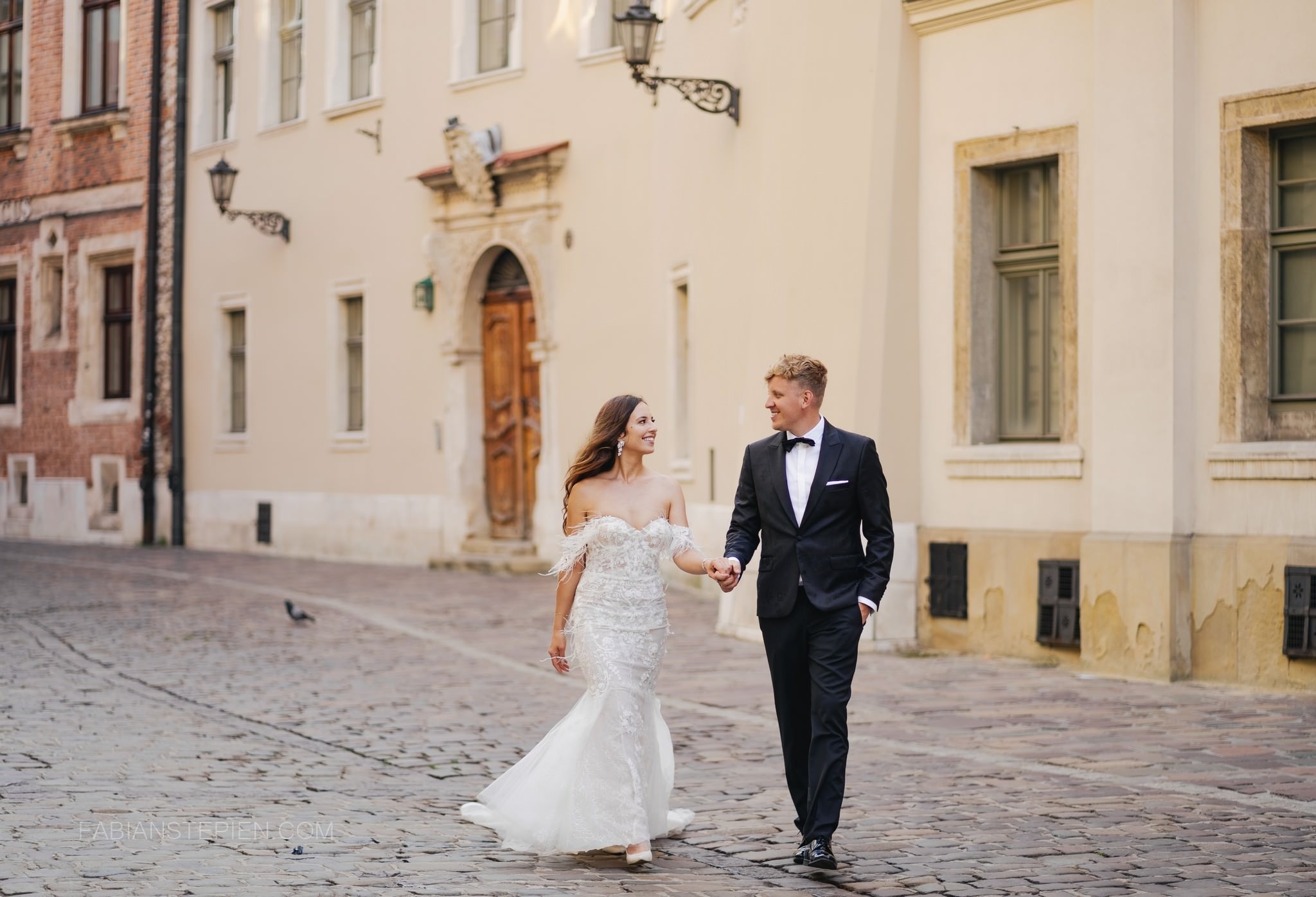 Uśmiechnięta para młoda idzie po brukowanej ulicy, trzymając się za ręce na tle architektury Starego Miasta. Ona w sukni z piórami, on w smokingu.