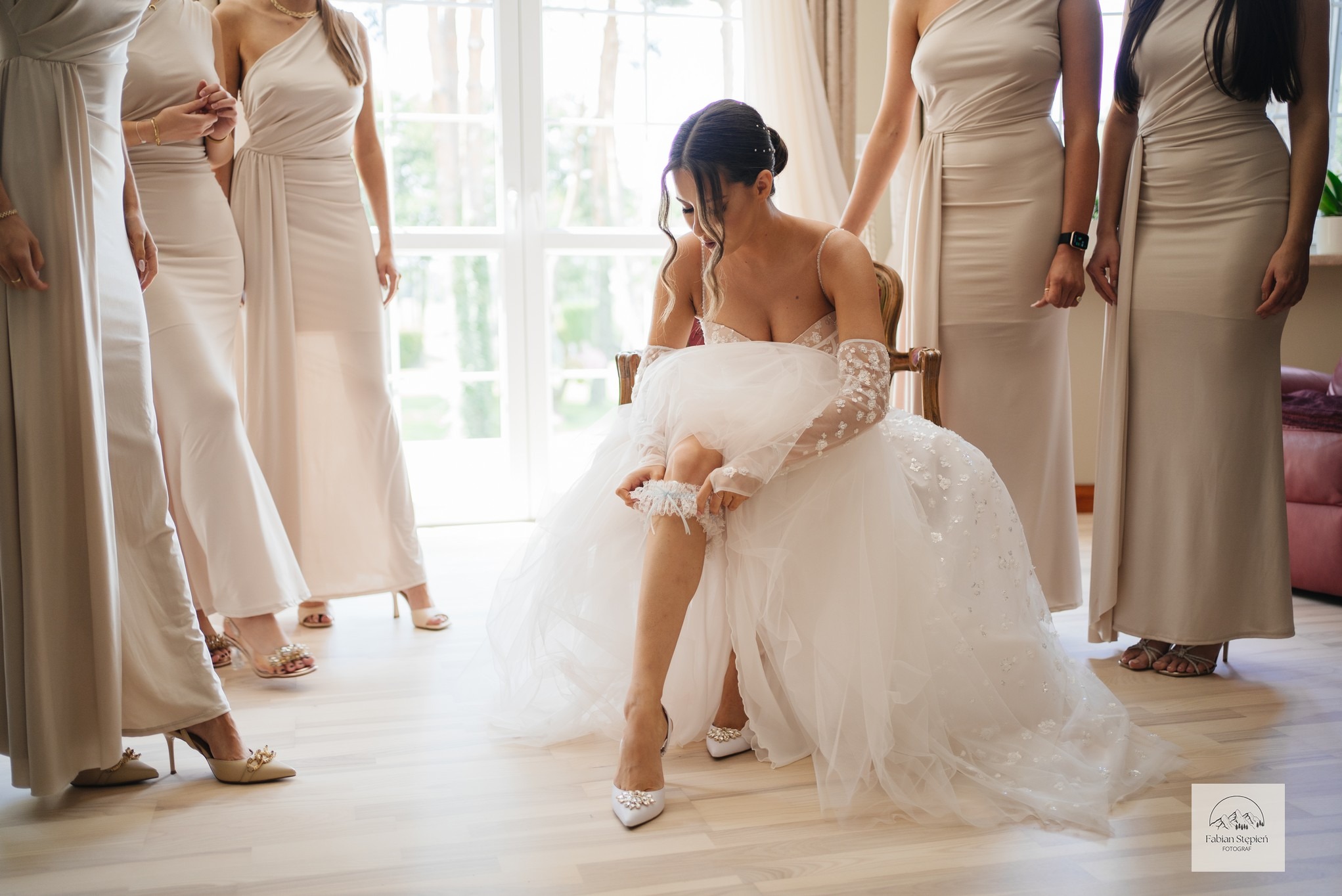 Panna młoda w sukni, siedząc na krześle, zakłada podwiązkę na tle druhen w beżowych sukniach. Jasne wnętrze, miękkie światło.
