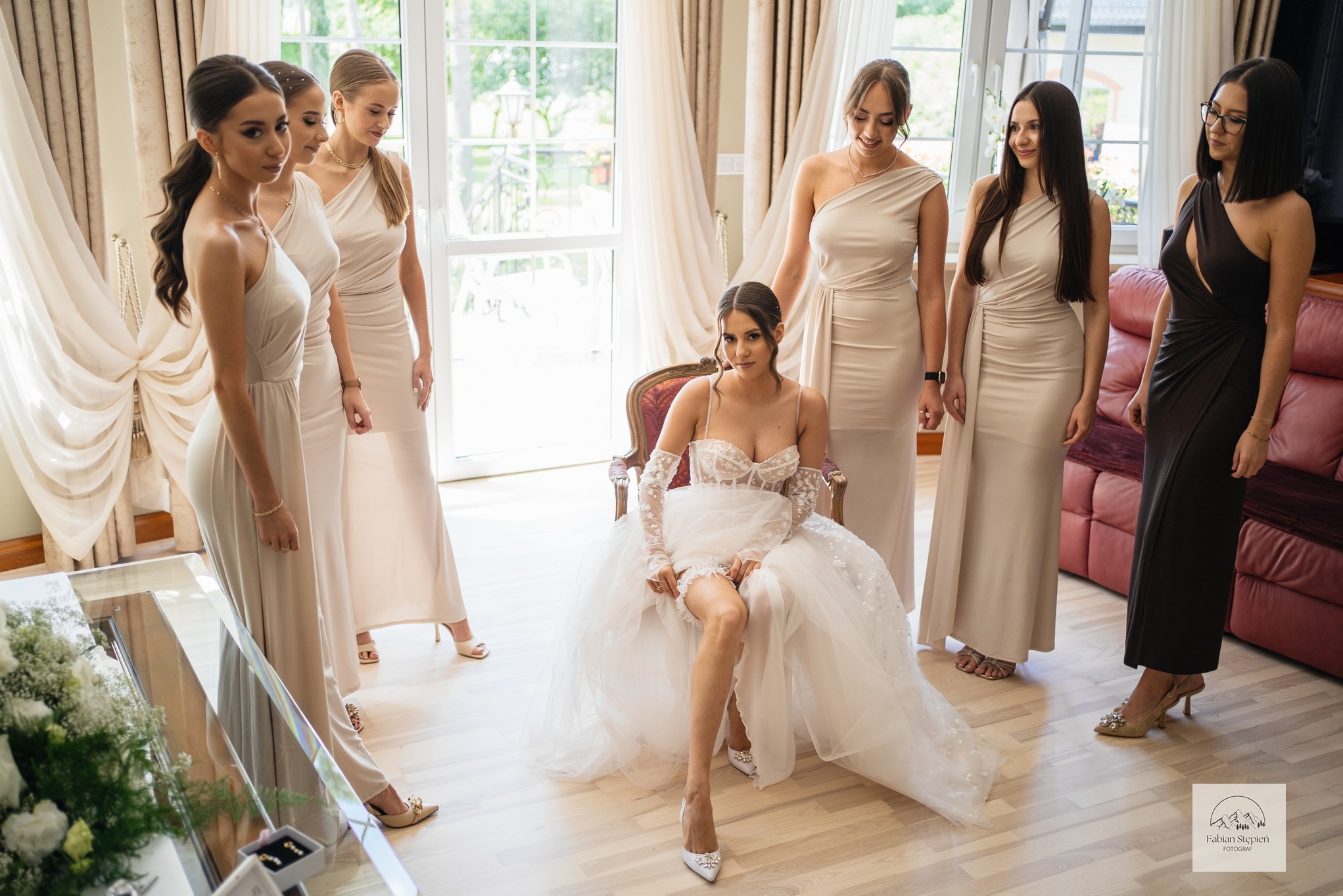 Panna młoda w koronkowej sukni siedzi na krześle, otoczona druhnami w beżowych sukienkach. Widoczne okno z firanami i kwiaty w tle. Przygotowania do ślubu.