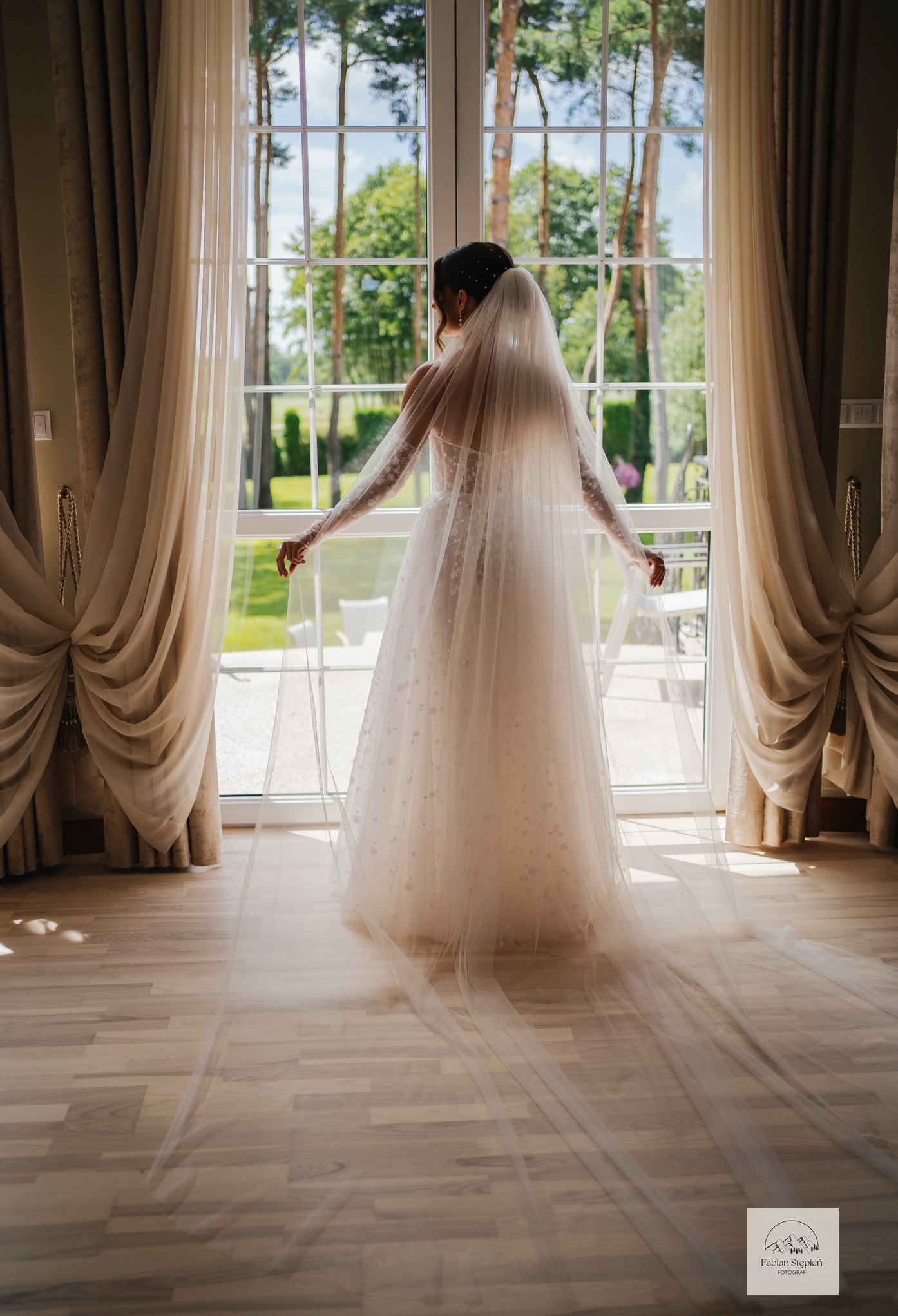 Panna młoda w koronkowej sukni, z długim welonem, stoi tyłem do okna z widokiem na zieleń. Delikatne światło podkreśla detale sukni i welonu.