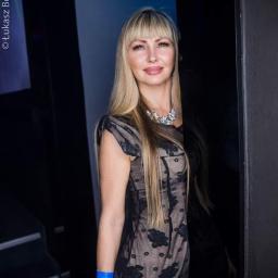 Viktoriiamodels - Portret blondwłosej kobiety w czarnej sukience z kwiatowym wzorem, stojącej na tle ciemnego wnętrza. Uwagę zwraca naszyjnik i bransoletka na ręce.