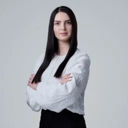 Portret kobiety o ciemnych włosach, ubrana w białą bluzkę, z założonymi rękami na neutralnym tle. Profesjonalny wizerunek.