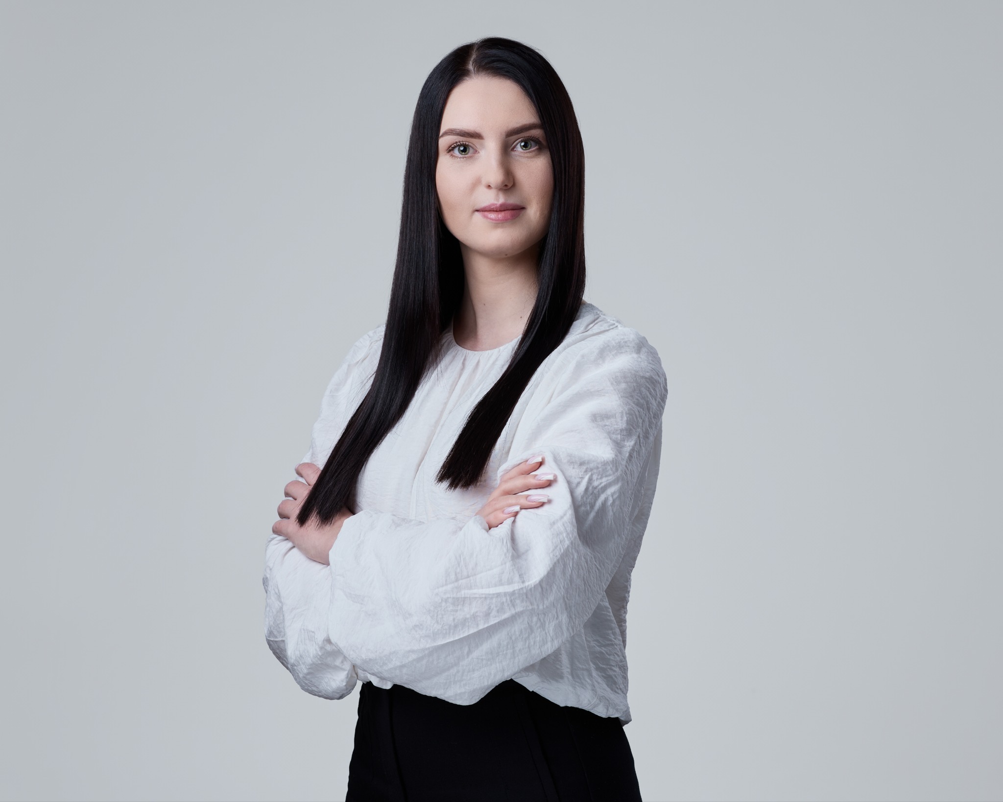 Portret kobiety o ciemnych włosach, ubrana w białą bluzkę, z założonymi rękami na neutralnym tle. Profesjonalny wizerunek.