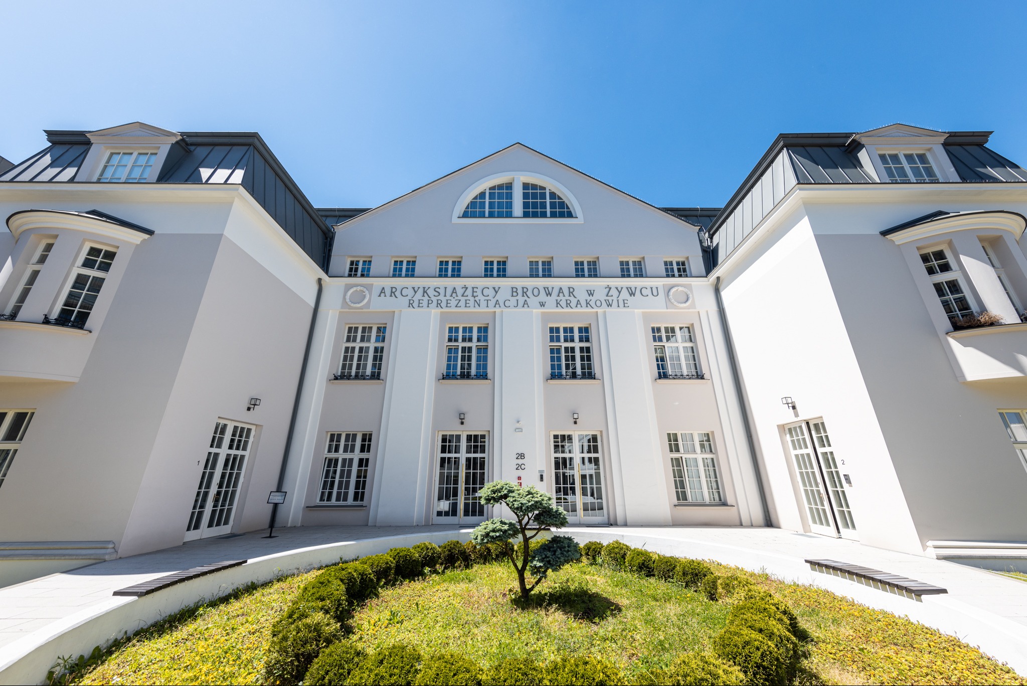 Elewacja budynku biurowego z napisem 'Arcyksiążęcy Browar w Żywcu Reprezentacja w Krakowie', zieleń przed wejściem, błękitne niebo w tle.