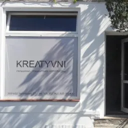 Fasada budynku z oknem, na którym widnieje szyld 'Kreatyvni Pracownia Projektowo Artystyczna', adres Jedności Narodowej 27 oraz numery telefonów, cień drzewa na ścianie, otwarte drzwi wejściowe.