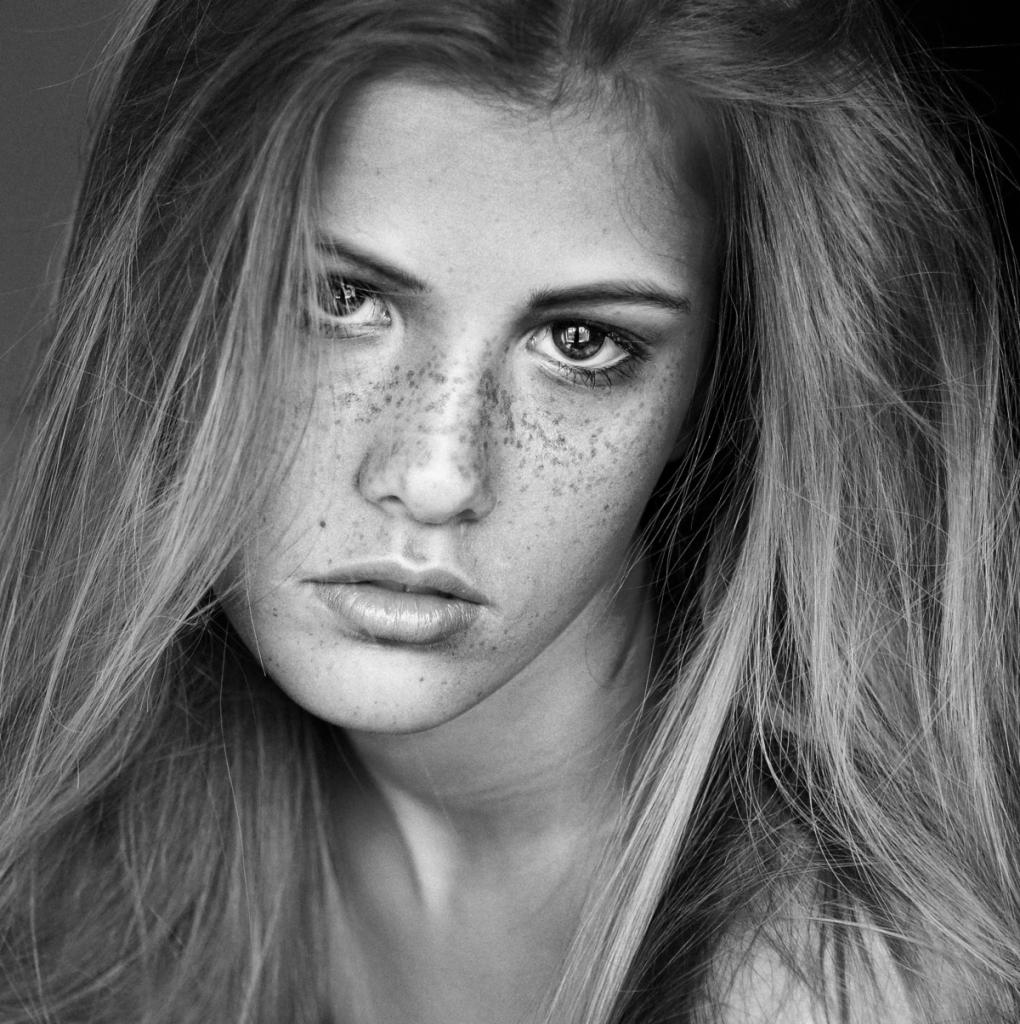 Czarno-biały portret kobiety z piegami, długimi włosami i intensywnym spojrzeniem.