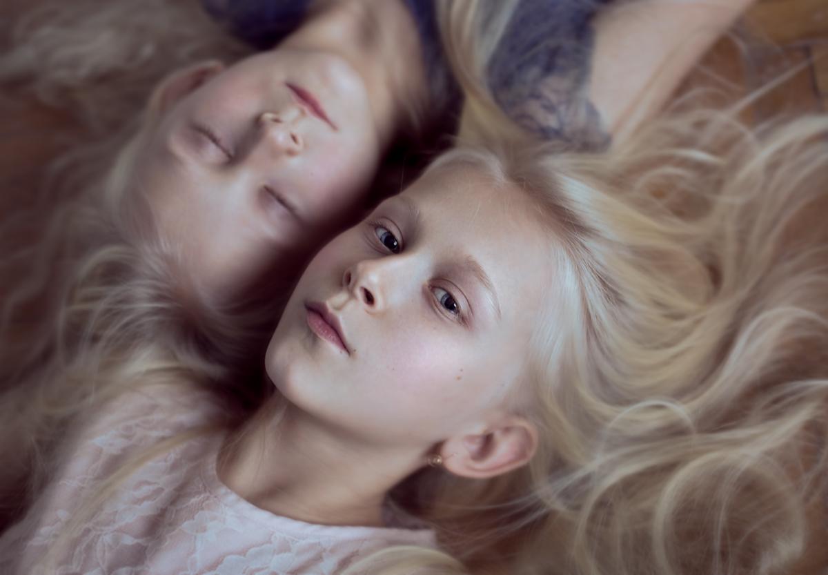 Portret dwóch jasnowłosych dziewczynek leżących obok siebie, jedna z zamkniętymi oczami, druga patrzy w obiektyw, na drewnianym tle.