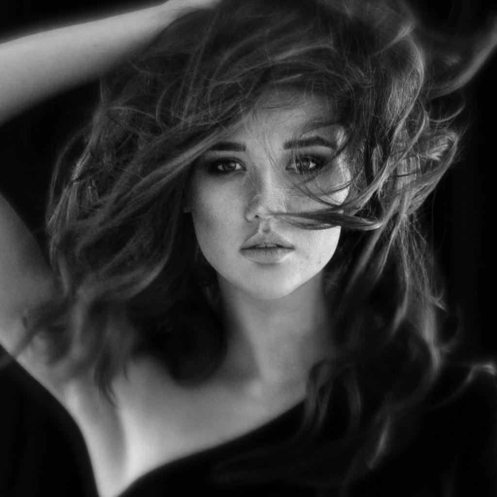 Czarno-biały portret kobiety z rozwianymi włosami, dłoń uniesiona nad głową, delikatne oświetlenie podkreśla rysy twarzy.