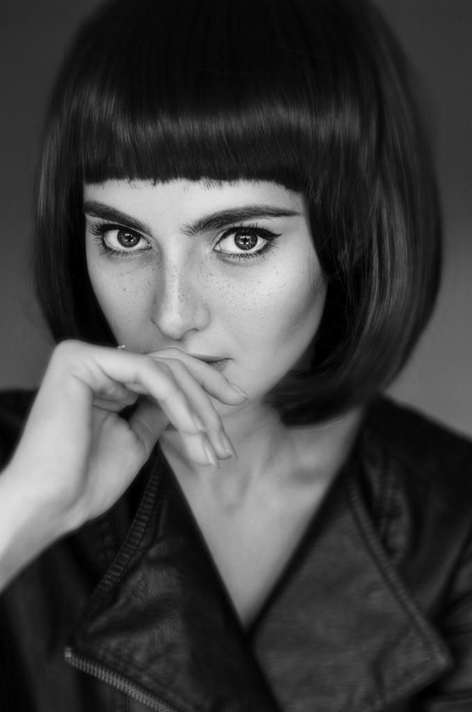 Czarno-biały portret kobiety z krótką, prostą grzywką, wyrazistym makijażem oczu i skórzaną kurtką, pozującej z dłonią przy ustach.