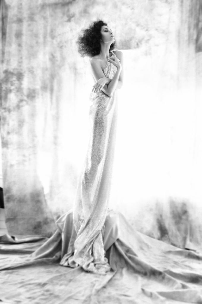 Czarno-białe zdjęcie portretowe kobiety w długiej, błyszczącej sukni, stojącej na drapowanym materiale na tle rozmytego tła.