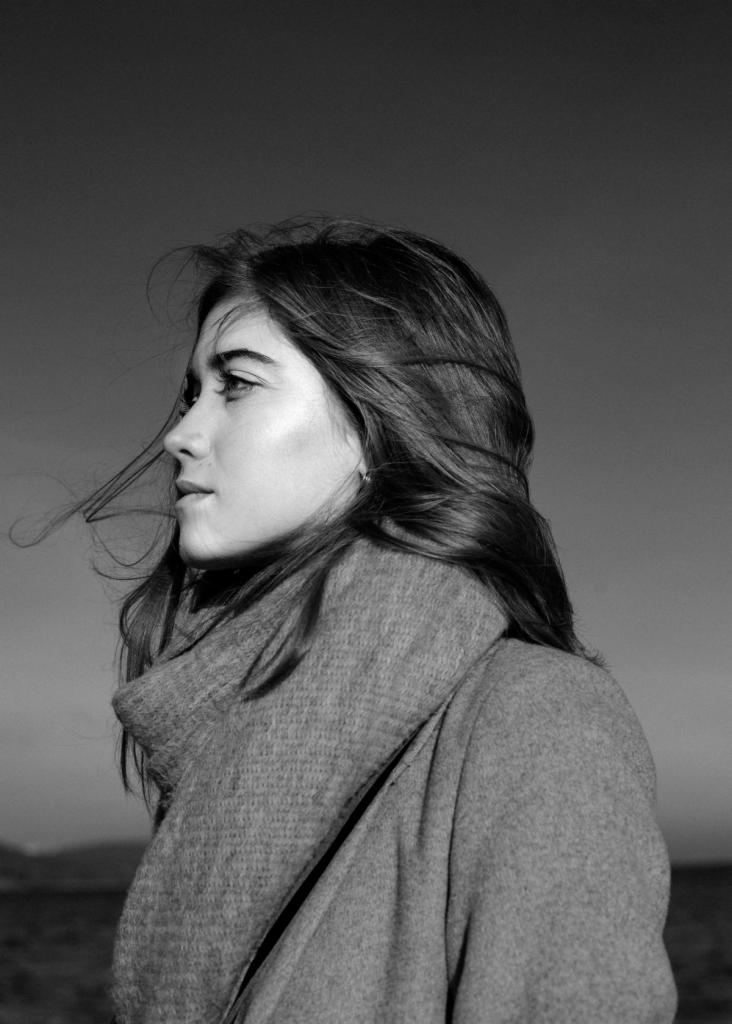 Czarno-biały portret profilowy kobiety z rozwianymi włosami na tle pochmurnego nieba i odległego krajobrazu.