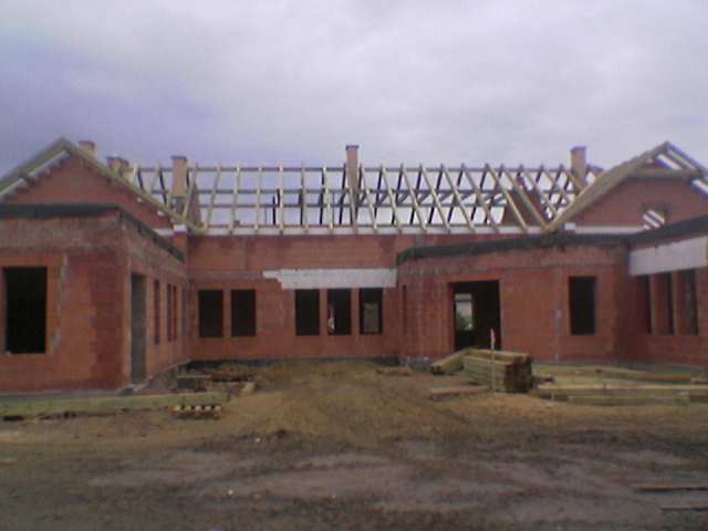 Budynek w trakcie budowy, z widoczną konstrukcją dachu z drewnianych belek, ściany z czerwonej cegły, teren budowy z ziemią i składowanym drewnem.