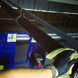 Adako - Ręka w czarnej rękawicy czyści zieloną ściereczką metalową konstrukcję pod sufitem, widoczny niebieski podnośnik z panelem sterowania.