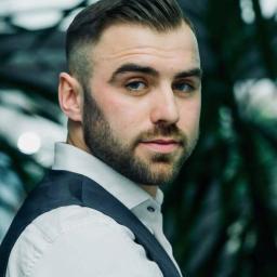 Adako - Mężczyzna z modną fryzurą fade i starannie przystrzyżoną brodą, ubrany w białą koszulę i kamizelkę, spogląda w bok, na tle zieleni. Logo salonu fryzjerskiego.