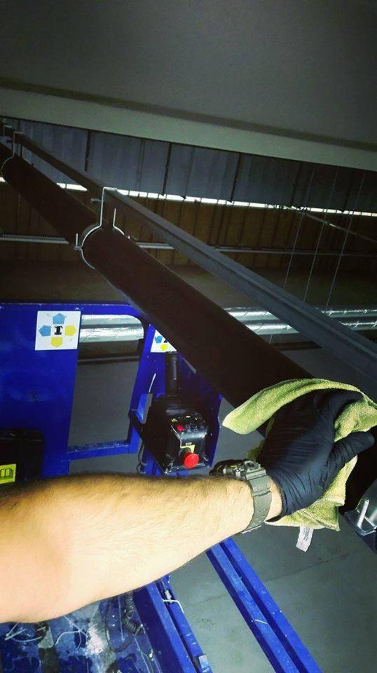 Ręka w czarnej rękawicy czyści zieloną ściereczką metalową konstrukcję pod sufitem, widoczny niebieski podnośnik z panelem sterowania.