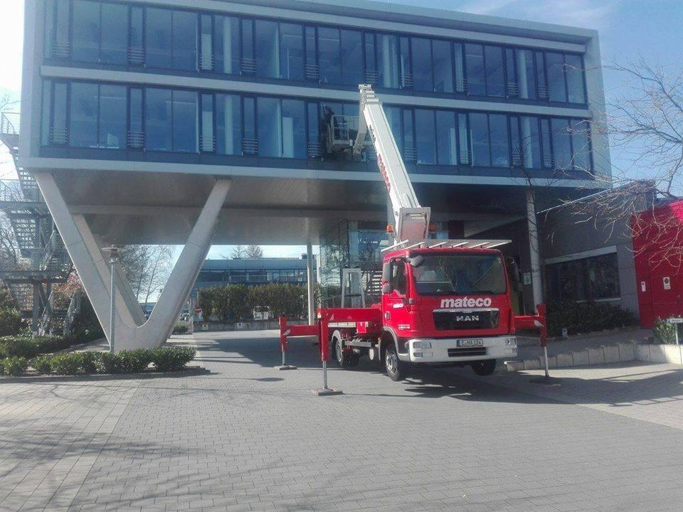 Czerwony podnośnik koszowy Mateco MAN ustawiony przed nowoczesnym biurowcem z dużą ilością szklanych okien; w koszu widoczna osoba podczas pracy.