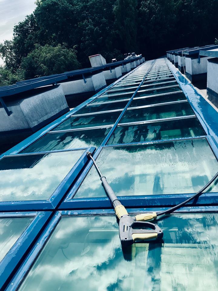 Mycie szklanego dachu budynku za pomocą myjki ciśnieniowej Karcher, widoczne odbicie chmur na umytej powierzchni.