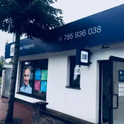 Fasada budynku z szyldem 'TAX CARE' i numerem telefonu, drzewo po lewej stronie, otwarte drzwi wejściowe.