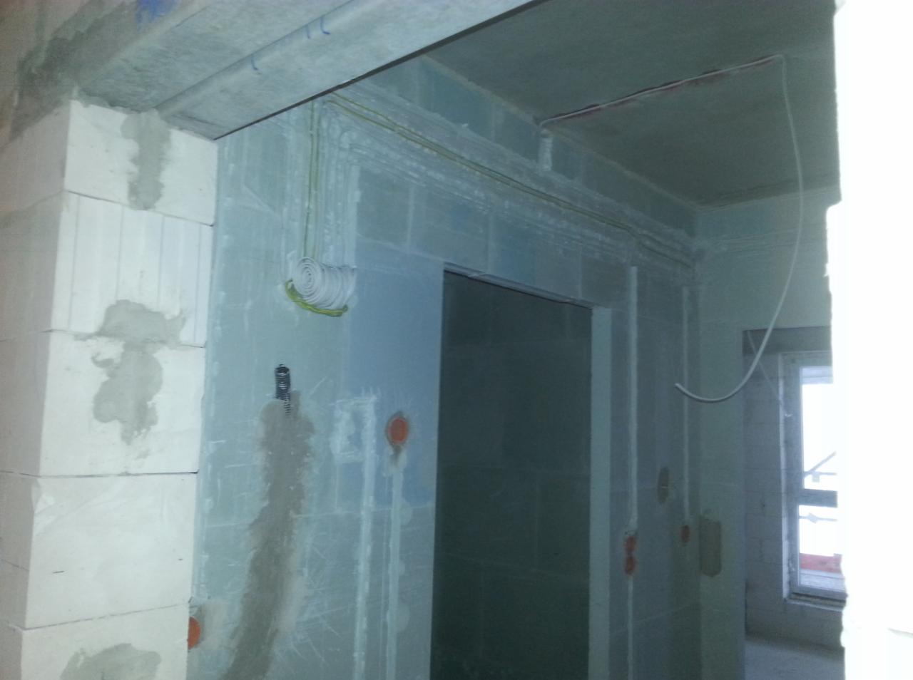 Surowe wnętrze w trakcie remontu: widoczne kable elektryczne prowadzone wzdłuż ścian z betonu komórkowego, zwinięty kabel w rolce, puszki instalacyjne oraz fragment okna.