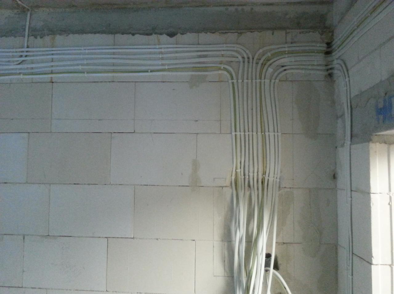 Białe kable elektryczne ułożone w rurkach na surowej ścianie z betonu komórkowego, widoczne łączenia i załamania kabli, surowy stan wykończenia wnętrza.