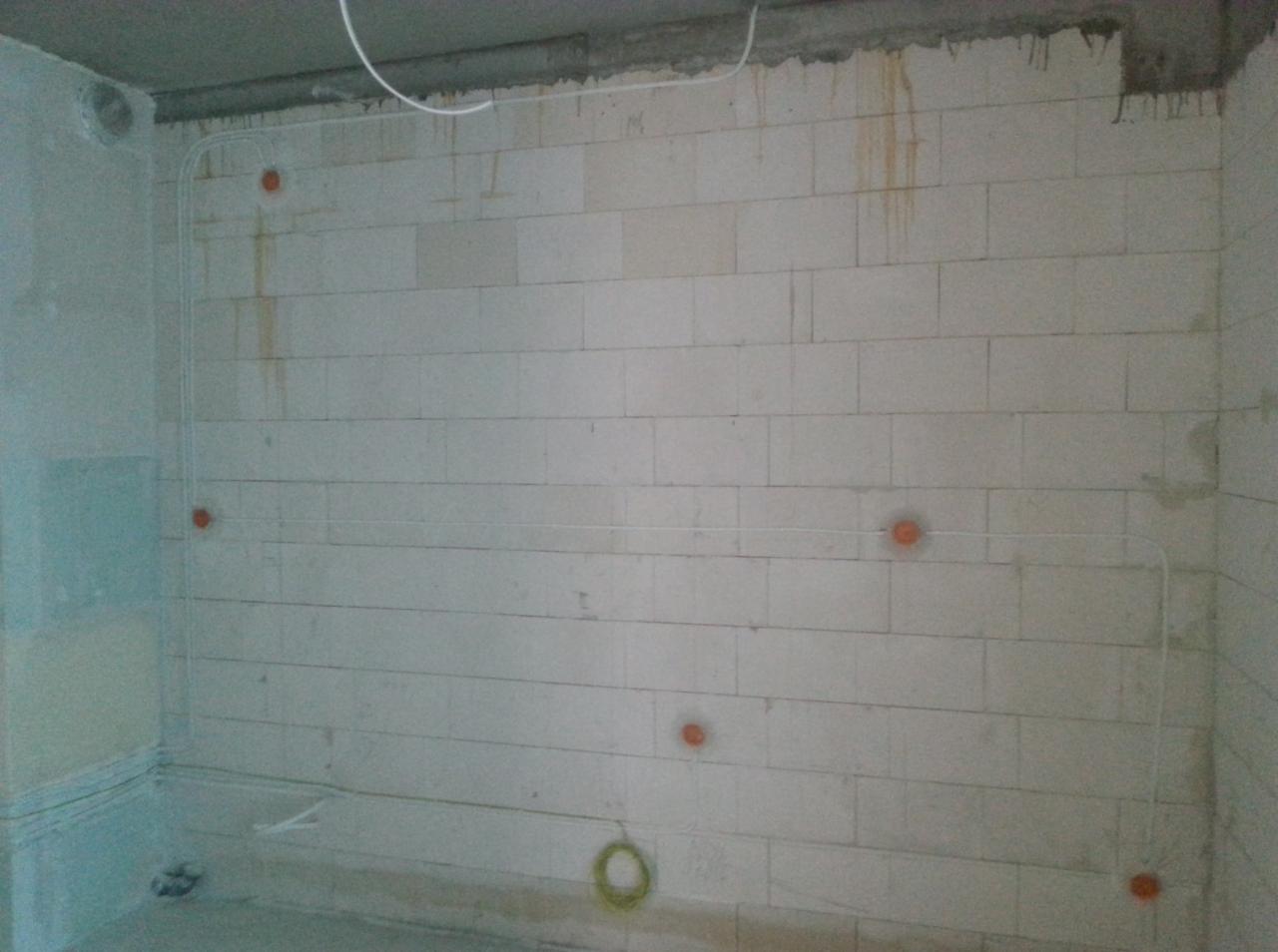 Surowa ściana z białych bloczków z widocznymi przewodami elektrycznymi poprowadzonymi w peszlach i puszkami instalacyjnymi w kolorze pomarańczowym, widok na wczesny etap prac.