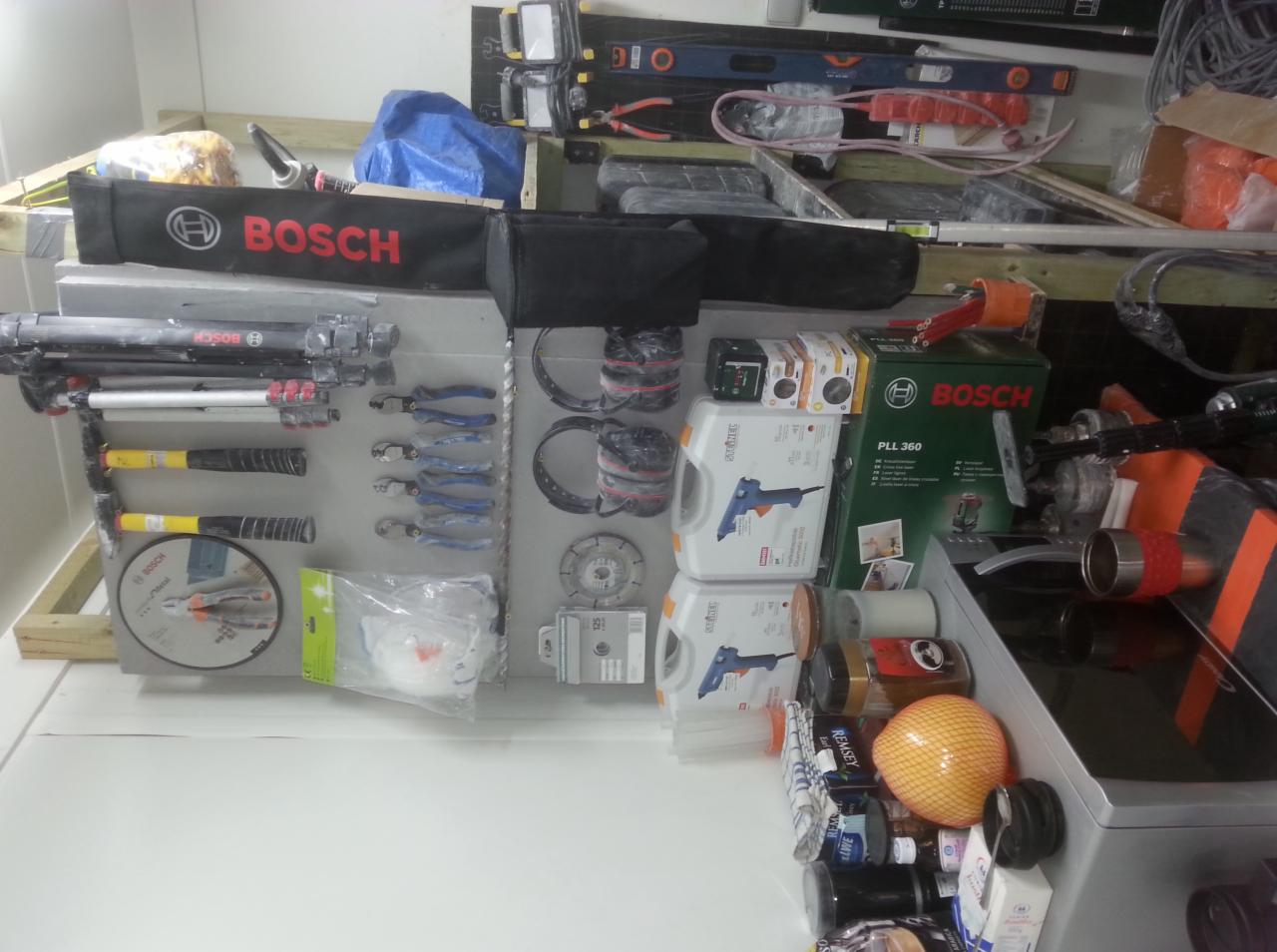 Zestaw narzędzi i akcesoriów elektrycznych marki Bosch, w tym poziomica laserowa, wiertła, młotki, szczypce i osłony słuchu, ułożone na półce w warsztacie.