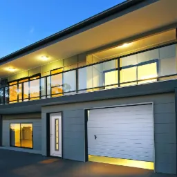 Dwukondygnacyjny dom z szarą elewacją, balkonem z przeszkloną balustradą, białymi drzwiami wejściowymi z przeszkleniami i segmentową bramą garażową z delikatnym oświetleniem od dołu.