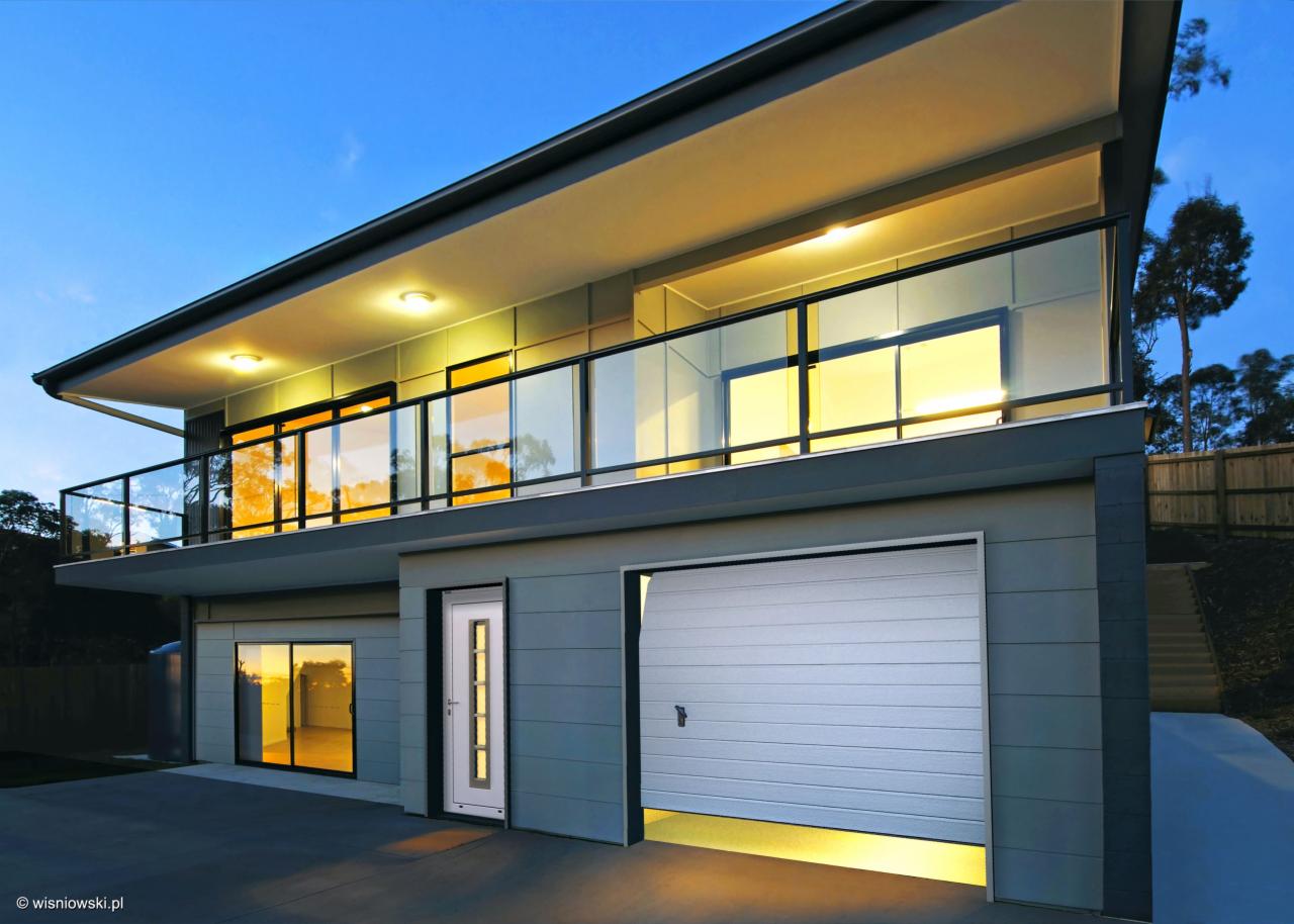 Dwukondygnacyjny dom z szarą elewacją, balkonem z przeszkloną balustradą, białymi drzwiami wejściowymi z przeszkleniami i segmentową bramą garażową z delikatnym oświetleniem od dołu.