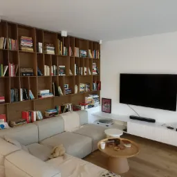 Jasny salon z narożną sofą, dużą drewnianą biblioteczką wypełnioną książkami i telewizorem na ścianie, okrągły stolik kawowy z drewnianym blatem i elementami dekoracyjnymi.