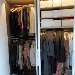 Wnętrze szafy wnękowej z otwartymi drzwiami, widoczne ubrania wiszące na wieszakach i białe pudełka do przechowywania na p&oacute;łkach.