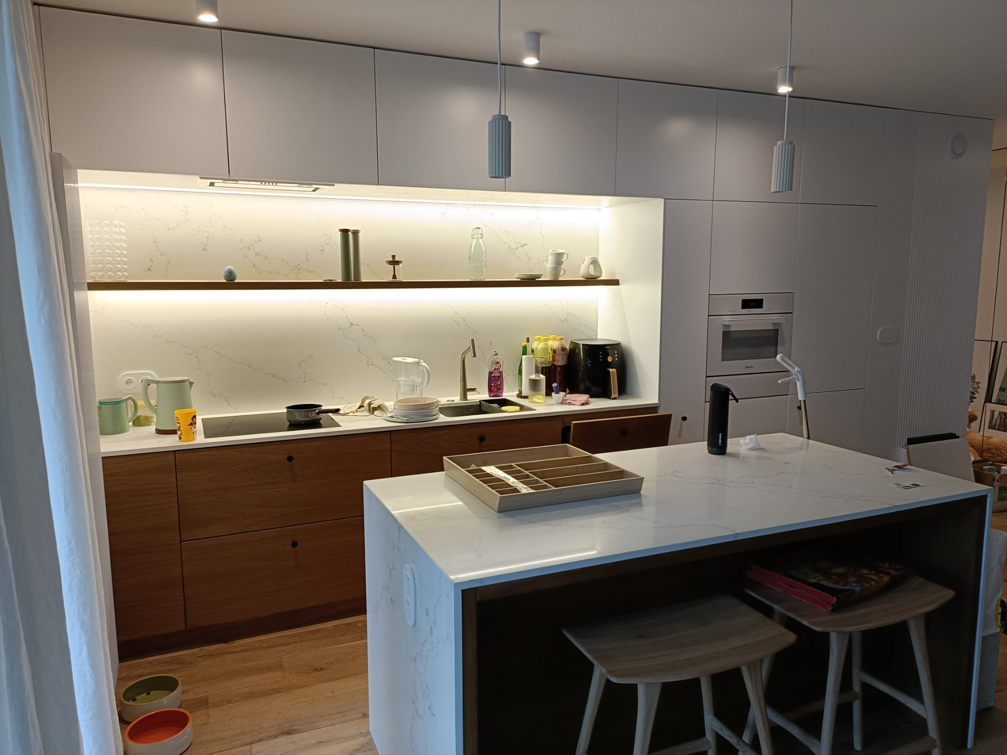 Jasna kuchnia z drewnianymi akcentami: zabudowa w bieli, drewniane fronty szafek dolnych, marmurowy blat wyspy z drewnianym obramowaniem, drewniane hokery, oświetlenie LED pod półkami...