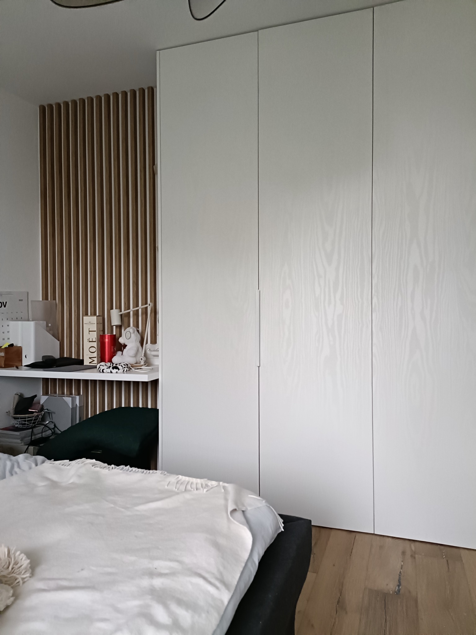 Biała, trzydrzwiowa szafa z drewnianą fakturą frontów, widoczna w sypialni obok łóżka i ażurowej ścianki z drewnianych listewek, z półką i dekoracjami.