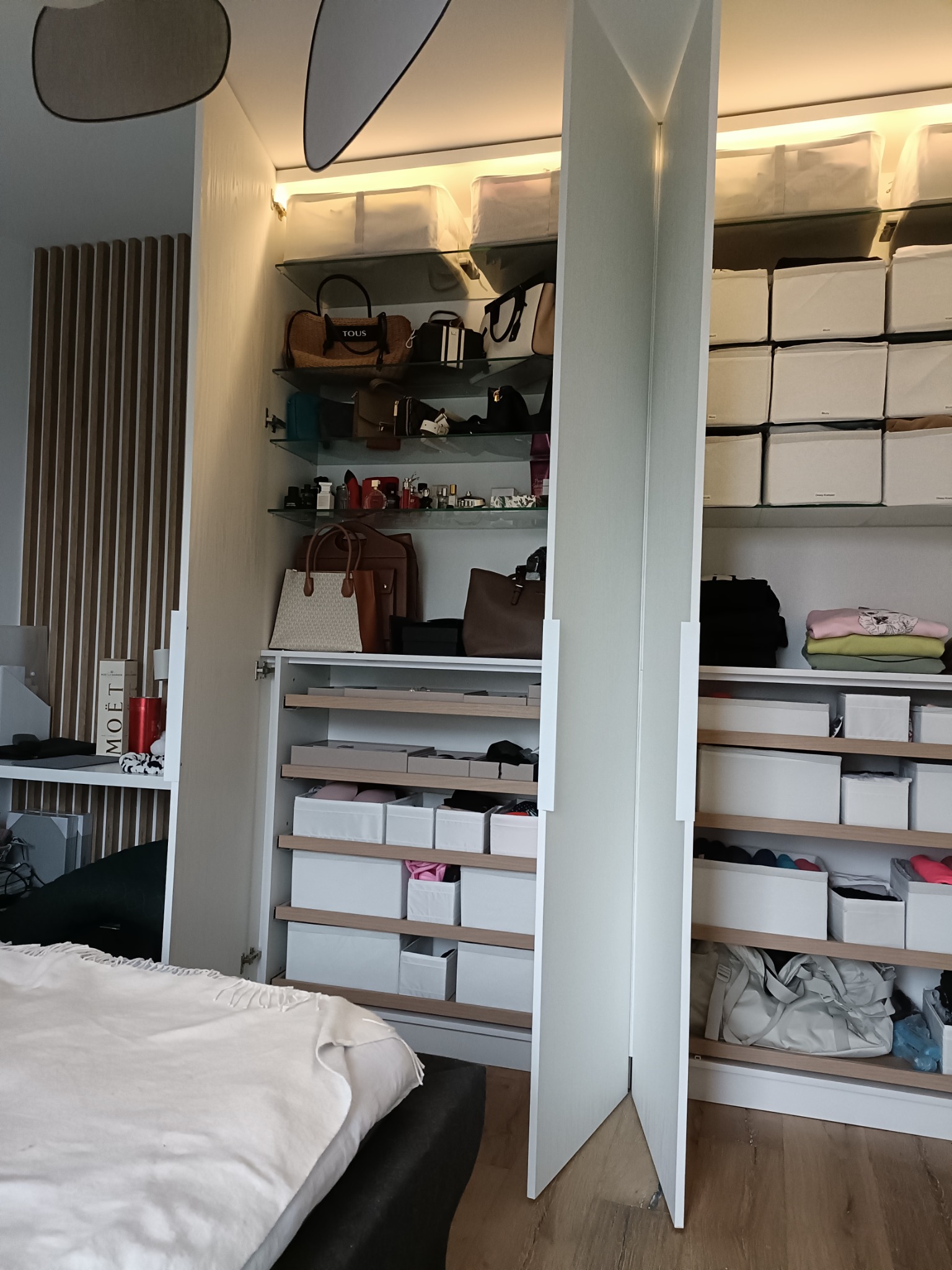 Otwarta szafa wnękowa z białymi frontami, szklanymi półkami i licznymi pudełkami do przechowywania, widoczne torebki, ubrania i kosmetyki, oświetlenie LED wewnątrz szafy.